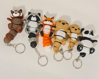 May include: A collection of handmade animal keychains. The keychains feature a raccoon, fox, otter, and panda bear. Each keychain is made of clay and has a silver keyring attached. The raccoon is brown and gray, the fox is orange and white.