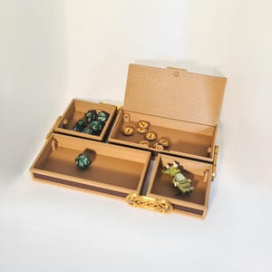 Pode incluir: Uma caixa multicompartimentos, de cor castanha clara, com tampa articulada, projetada para armazenar acessórios de jogos. Os compartimentos contêm dados, moedas e uma pequena figura. A caixa tem um fecho e acabamento decorativos dourados.