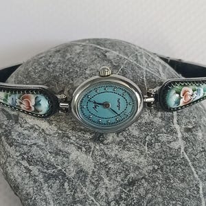 May include: A silver-tone oval wristwatch with a light blue dial featuring dark blue hands and a dotted border. The bracelet links are decorated with hand-painted floral motifs in shades of pink, blue, and green on a white background.