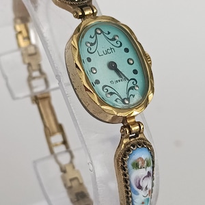 Peut inclure: Montre-bracelet Luch dorée vintage avec un cadran ovale bleu clair. La montre porte l'inscription "Luch" et "15 jewels". Le bracelet est orné d'émail floral bleu, blanc et violet.