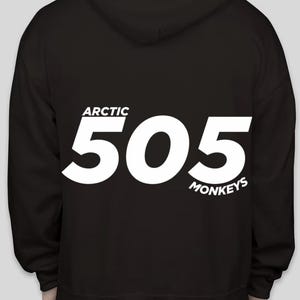 Puede incluir: Sudadera con capucha negra con el texto blanco "ARCTIC 505 MONKEYS". La sudadera tiene capucha y mangas largas. El texto está centrado en la parte trasera.