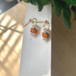 May include: A pair of dangle earrings featuring white coffee cup charms with gold-toned hoops. Each cup has a textured brown sleeve adorned with a small orange pumpkin accent. The earrings are suspended from gold-toned ball-post earring findings.