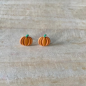 May include: Two orange pumpkin-shaped earrings with green stems. The earrings are set against a light-colored wooden background. These earrings are perfect for fall and Halloween.