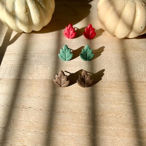 May include: A collection of leaf-shaped stud earrings in red, green, and brown. The earrings are displayed on a wooden surface with two white pumpkins in the background. The earrings are small and delicate, perfect for fall.