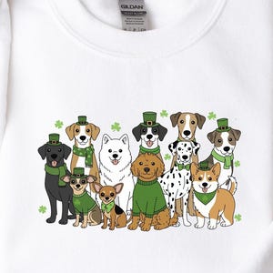 Peut inclure: Sweat-shirt blanc avec un motif de différentes races de chiens, chacun portant un chapeau et une écharpe verts avec des détails de trèfles. Le mot "White" est imprimé dans le coin supérieur droit. Le sweat-shirt est en matière douce.