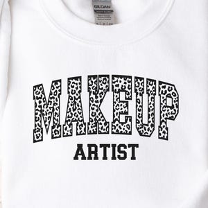 May include: White crewneck sweatshirt with the words "MAKEUP ARTIST" in a black leopard print design. The sweatshirt is made of a soft material and is perfect for makeup artists.