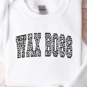 Peut inclure: Sweat-shirt blanc avec l'inscription "TAX BOSS" en motif léopard noir et blanc. Le vêtement est posé sur une surface en bois.