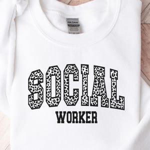 Puede incluir: Sudadera blanca con la frase "SOCIAL WORKER" en negro, con letras con estampado de leopardo. La sudadera está hecha de un material suave y se muestra sobre una superficie de madera.