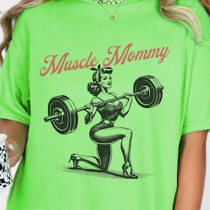 May include: Green t-shirt with a black and white illustration of a woman lifting weights. The text "Muscle Mommy" is printed in red on the shirt.