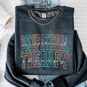 Könnte beinhalten: Dunkelgraues Sweatshirt mit dem Schriftzug "OCCUPATIONAL THERAPY" in einer bunten Retro-Schriftart. Das Sweatshirt wird mit einer blauen Jeans, einer goldenen Kette und weißen High-Top-Sneakern kombiniert. Der Text "Dark Heather Grey" befindet sich oben rechts.