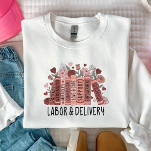 May include: A white sweatshirt with the words "LABOR & DELIVERY" in black. The design includes a stack of books with words like "SUPPORT" and "EMPOWERMENT", surrounded by flowers and hearts. A pink cap and blue jeans are also in the image.