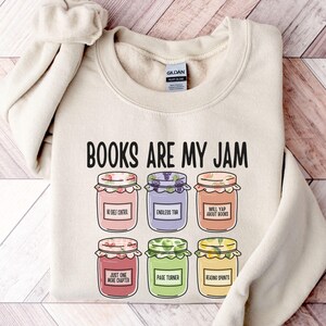 以下が含まれることがあります： 「BOOKS ARE MY JAM」の文字と、6つのイラスト入りのジャム瓶が描かれたクリーム色のスウェットシャツ。「No Self Control」や「Endless Tea」など、それぞれの瓶には異なる色とラベルが施されています。柔らかい素材で作られています。