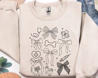 May include: A sand-colored crewneck sweatshirt featuring a black line art graphic print. The design includes various bows, a stethoscope, a bone, a bandage, a syringe, a heart with a cross, and medical gloves arranged in a central pattern.