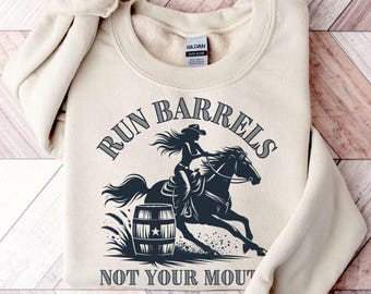 May include: A sand-colored crewneck sweatshirt featuring a dark graphic of a person riding a horse toward a barrel. The text "RUN BARRELS" is arched above the graphic, and the text "NOT YOUR MOUTH" is printed below the barrel illustration.