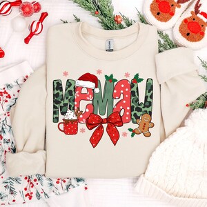 May include: A cream-colored crewneck sweatshirt featuring the word MEMAW in a festive graphic print. The letters are decorated with green leopard print, red polka dots, a Santa hat, a red bow, a gingerbread man, and a mug of hot cocoa.