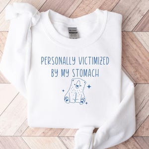 以下が含まれることがあります： 胸元に青い文字で「PERSONALLY VICTIMIZED BY MY STOMACH」とプリントされた、白い長袖のクルーネックトレーナー。文字の下には、4つの小さな星に囲まれた、座っているクマの青い線画イラストが描かれています。