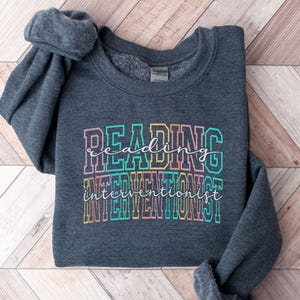 Può includere: Una felpa girocollo grigio scuro mélange con la scritta "READING INTERVENTIONIST" in un font a blocchi multicolore. La parola "reading" è in carattere corsivo bianco, e "interventionist" è in carattere corsivo bianco sovrapposto al testo a blocchi.
