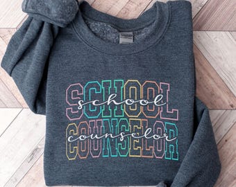 May include: A dark heather gray crewneck sweatshirt featuring the text "SCHOOL COUNSELOR" in a multi-colored, outlined collegiate font. The words "school" and "counselor" are also written in a white cursive script layered over the block letters.