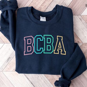 May include: A navy blue crewneck sweatshirt featuring the letters BCBA printed in a collegiate font across the chest. The letters are outlined in a dark navy color, with the B in pink, the C and B in teal, and the A in yellow. The text Navy is visible.