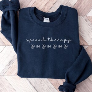 May include: A navy blue crewneck sweatshirt featuring the white text speech therapy in a cursive script font, positioned above a horizontal row of seven small, white, outlined bow illustrations centered on the front of the garment.