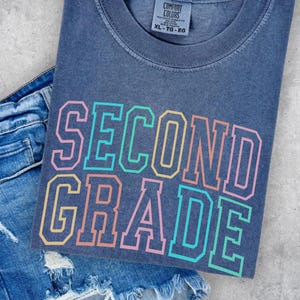 May include: A slate blue crew neck t-shirt with a vintage wash finish. The shirt features the text SECOND GRADE printed in a bold, collegiate-style font with an outline design in alternating colors of pink, teal, yellow, and orange.
