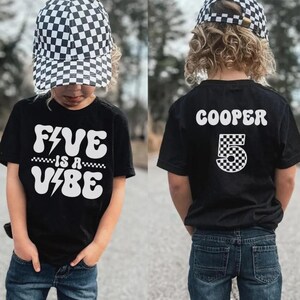May include: A black t-shirt featuring white text that reads FIVE IS A VIBE with lightning bolt accents on the front and the name COOPER above a large number 5 with a checkered pattern on the back, paired with a black and white checkered baseball cap.