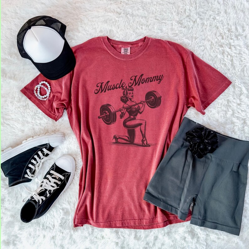 May include: Red t-shirt with a vintage style illustration of a woman lifting a barbell. The text "Muscle Mommy" is printed on the shirt.
