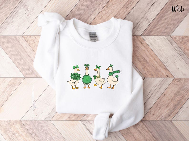 May include: White sweatshirt with a St. Patrick's Day design. Four cartoon geese are depicted, each wearing green accessories such as shamrocks, hats, and scarves. The word "White" is printed in the upper right corner.