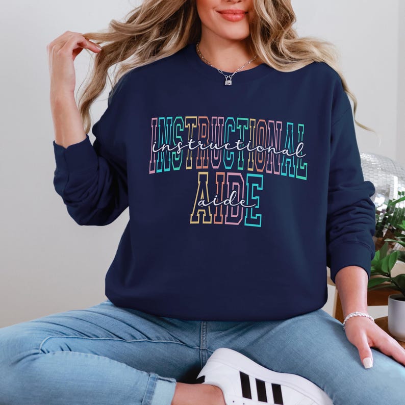 May include: Navy blue sweatshirt with the words "INSTRUCTIONAL AIDE" in a colorful, outlined font. The text is in a gradient of pink, blue, and yellow. The sweatshirt is a crew neck style.