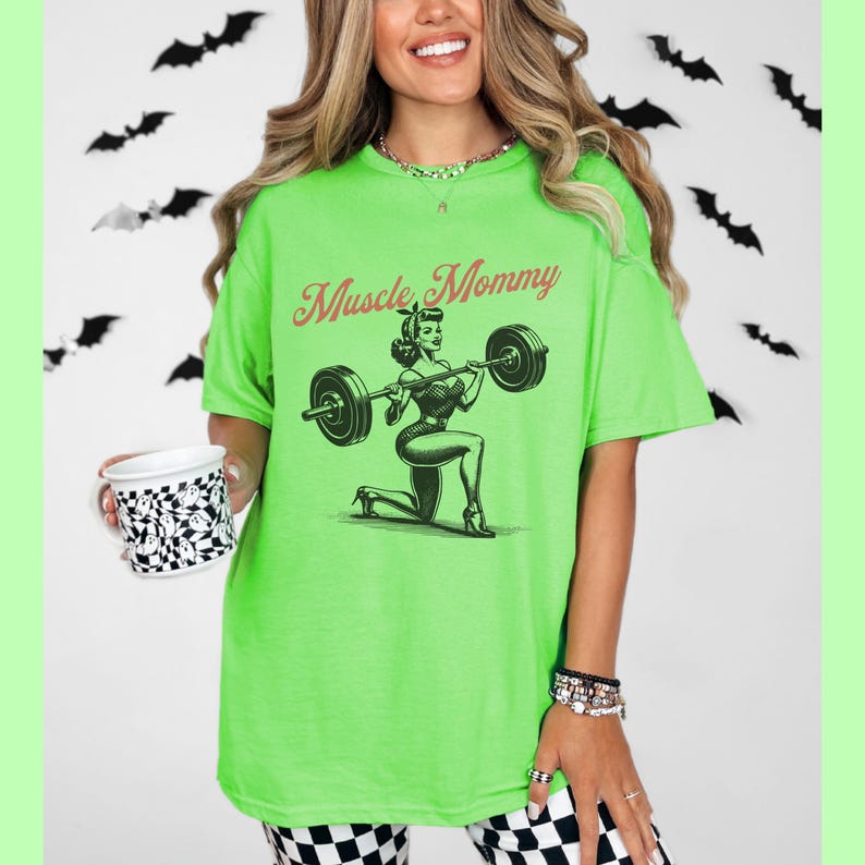 May include: Green t-shirt with a black and white illustration of a woman lifting weights. The text "Muscle Mommy" is printed in red on the shirt.