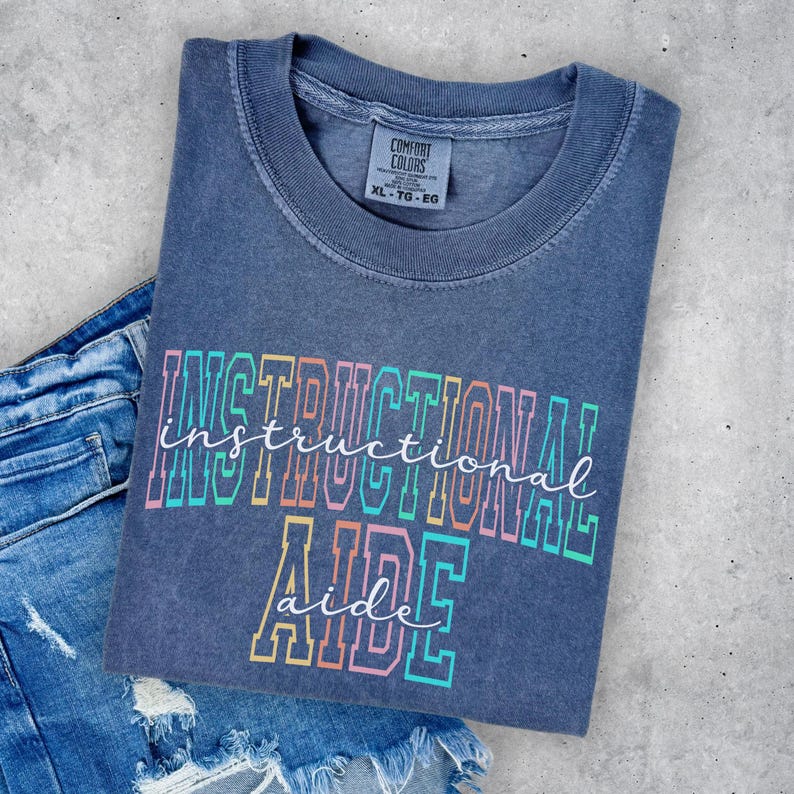 May include: A blue Comfort Colors t-shirt with the words "INSTRUCTIONAL AIDE" in a colorful, outlined font. The shirt is folded and placed on top of a pair of denim shorts. The shirt has a crew neck and short sleeves.