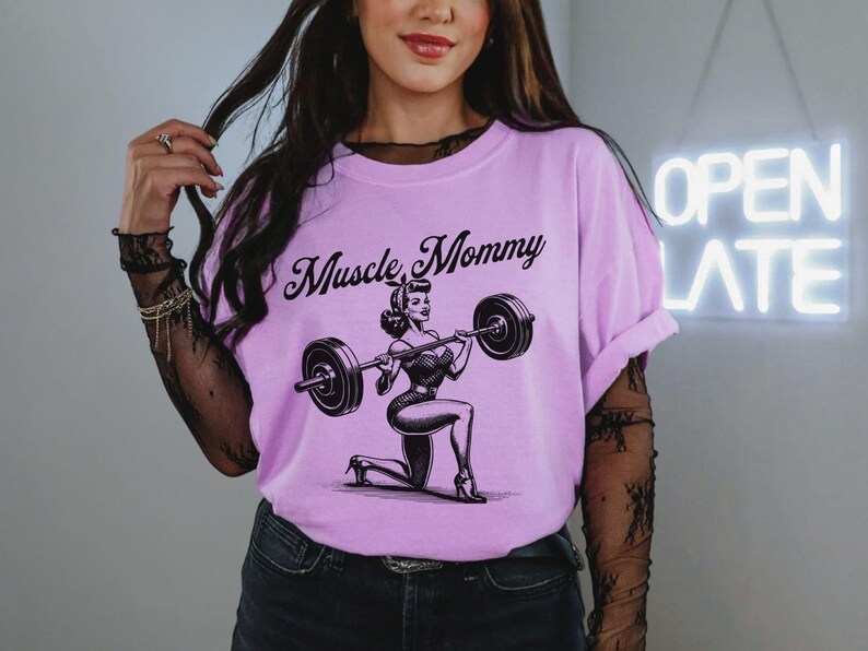 May include: Pink t-shirt with a black and white illustration of a woman lifting a barbell. The text "Muscle Mommy" is printed on the shirt.