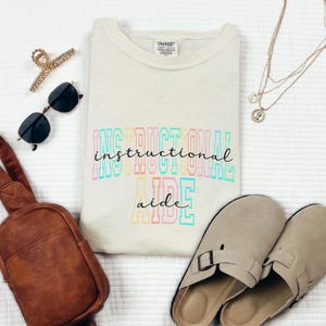 May include: Cream-colored t-shirt with the words "INSTRUCTIONAL AIDE" in colorful block letters. The image also includes sunglasses, a brown bag, a gold hair clip, a layered necklace, and beige slip-on shoes.