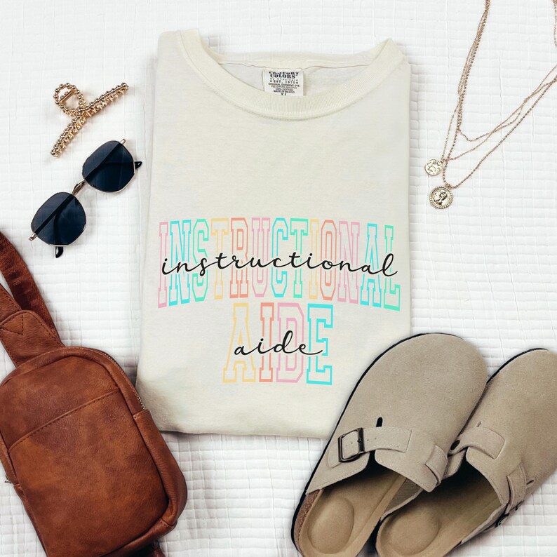 May include: Cream-colored t-shirt with the words "INSTRUCTIONAL AIDE" in colorful block letters. The image also includes sunglasses, a brown bag, a gold hair clip, a layered necklace, and beige slip-on shoes.