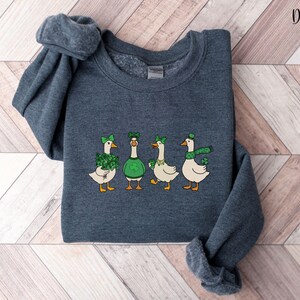 May include: Dark heather grey sweatshirt with a St. Patrick's Day themed design. The design features four geese, each wearing green accessories such as hats, bows, and scarves. The text "Dark Heather" is in the upper right corner.