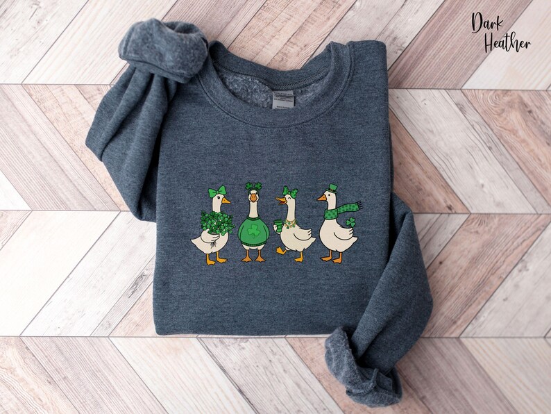 May include: Dark heather grey sweatshirt with a St. Patrick's Day themed design. The design features four geese, each wearing green accessories such as hats, bows, and scarves. The text "Dark Heather" is in the upper right corner.