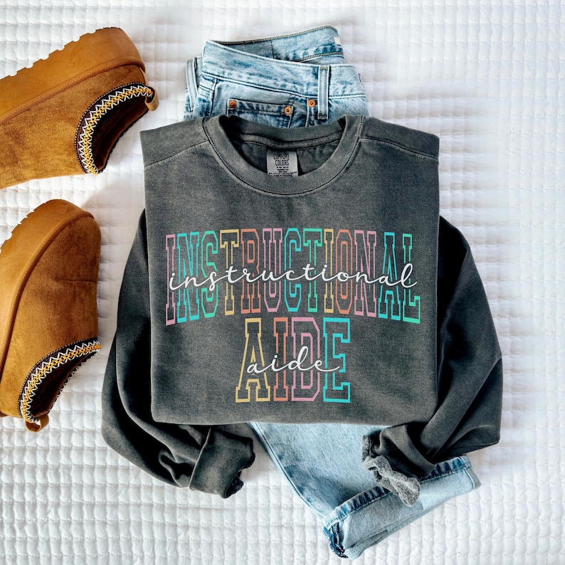 May include: A dark gray sweatshirt with the words "INSTRUCTIONAL AIDE" in a colorful, layered font. The sweatshirt is paired with light blue jeans and brown suede slippers with decorative stitching.