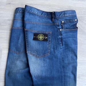 May include: Blue denim jeans with a classic five-pocket design. The jeans feature a dark wash with subtle fading and a small patch on the back pocket. The patch has a circular logo with a compass design.