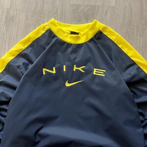 May include: Navy blue pullover with yellow accents and the Nike logo in yellow. The top has a crew neck and yellow stripes down the sleeves. The Nike swoosh is below the logo.