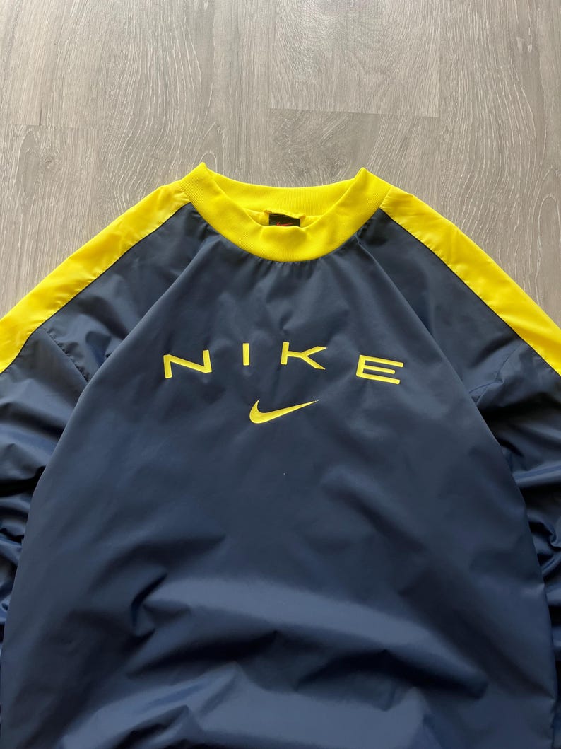 May include: Navy blue pullover with yellow accents and the Nike logo in yellow. The top has a crew neck and yellow stripes down the sleeves. The Nike swoosh is below the logo.