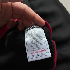 May include: Black sweatshirt with red trim. A tag with the text "F8TWA" and fabric composition details is visible. The tag also includes the text "KEEP AWAY FROM FIRE" and "MADE IN MALAYSIA".