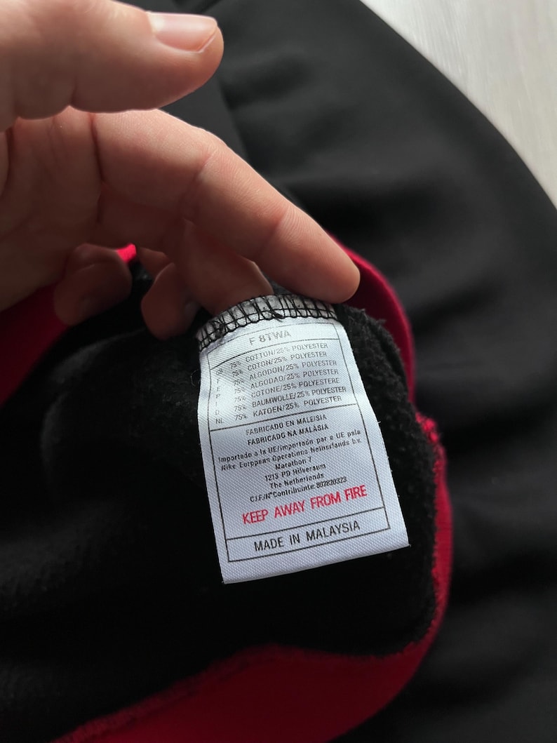 May include: Black sweatshirt with red trim. A tag with the text "F8TWA" and fabric composition details is visible. The tag also includes the text "KEEP AWAY FROM FIRE" and "MADE IN MALAYSIA".