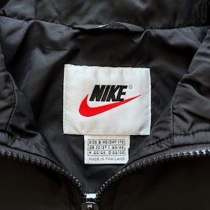 May include: Black Nike jacket with a blue collar. The Nike logo is prominently displayed on a white tag. The jacket has a zipper closure and a tag that says "MADE IN THAILAND".