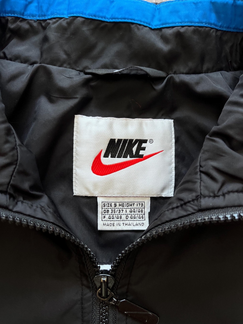 May include: Black Nike jacket with a blue collar. The Nike logo is prominently displayed on a white tag. The jacket has a zipper closure and a tag that says "MADE IN THAILAND".