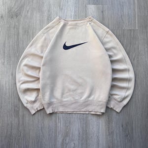 May include: A beige crew neck sweatshirt with a navy blue Nike swoosh logo on the chest. The sweatshirt has ribbed cuffs and hem. The garment is made of a soft material.