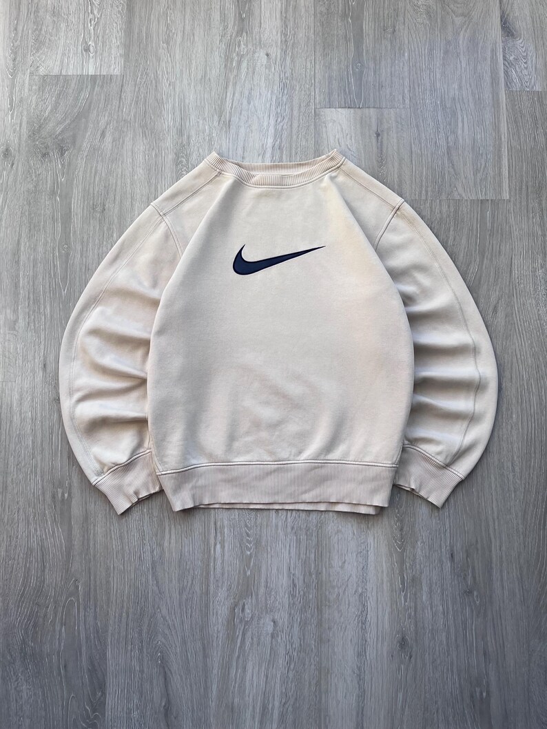 May include: A beige crew neck sweatshirt with a navy blue Nike swoosh logo on the chest. The sweatshirt has ribbed cuffs and hem. The garment is made of a soft material.