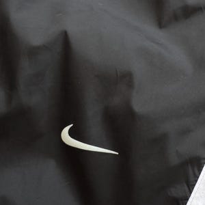 Peut inclure: Veste de sport noire avec le logo Nike blanc sur le devant. La veste a une bande blanche le long du bord. Le mat&eacute;riau semble &ecirc;tre un tissu l&eacute;ger et r&eacute;sistant &agrave; l'eau.