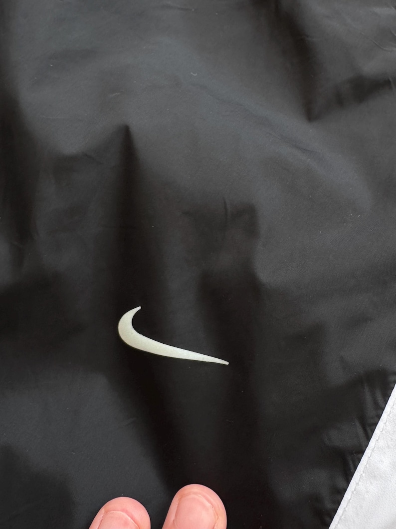 Peut inclure: Veste de sport noire avec le logo Nike blanc sur le devant. La veste a une bande blanche le long du bord. Le mat&eacute;riau semble &ecirc;tre un tissu l&eacute;ger et r&eacute;sistant &agrave; l'eau.