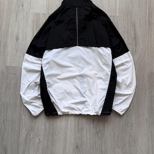 Peut inclure: Veste coupe-vent noire et blanche. La partie sup&eacute;rieure et les panneaux lat&eacute;raux sont noirs, tandis que l'avant et les manches sont blancs. La veste a un col montant et un cordon de serrage &agrave; la taille. Une fine bande argent&eacute;e descend le dos.