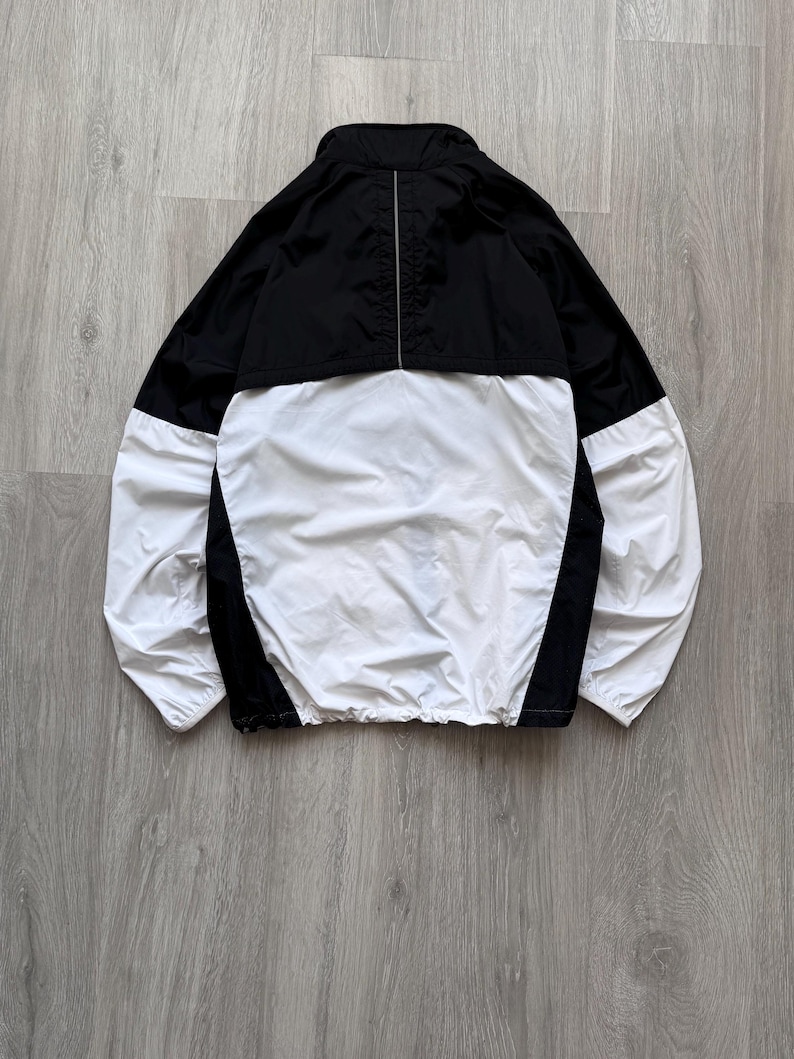 Peut inclure: Veste coupe-vent noire et blanche. La partie sup&eacute;rieure et les panneaux lat&eacute;raux sont noirs, tandis que l'avant et les manches sont blancs. La veste a un col montant et un cordon de serrage &agrave; la taille. Une fine bande argent&eacute;e descend le dos.